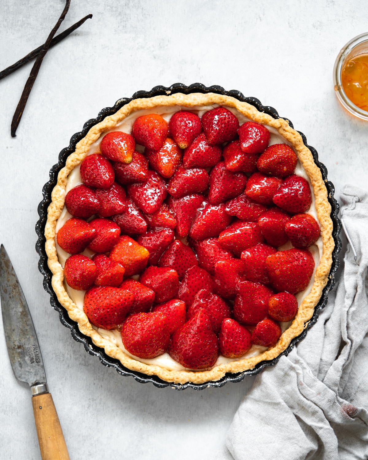 top down view of french strawberry tart with grey linen, a knife and vanilla pods scattered around it.