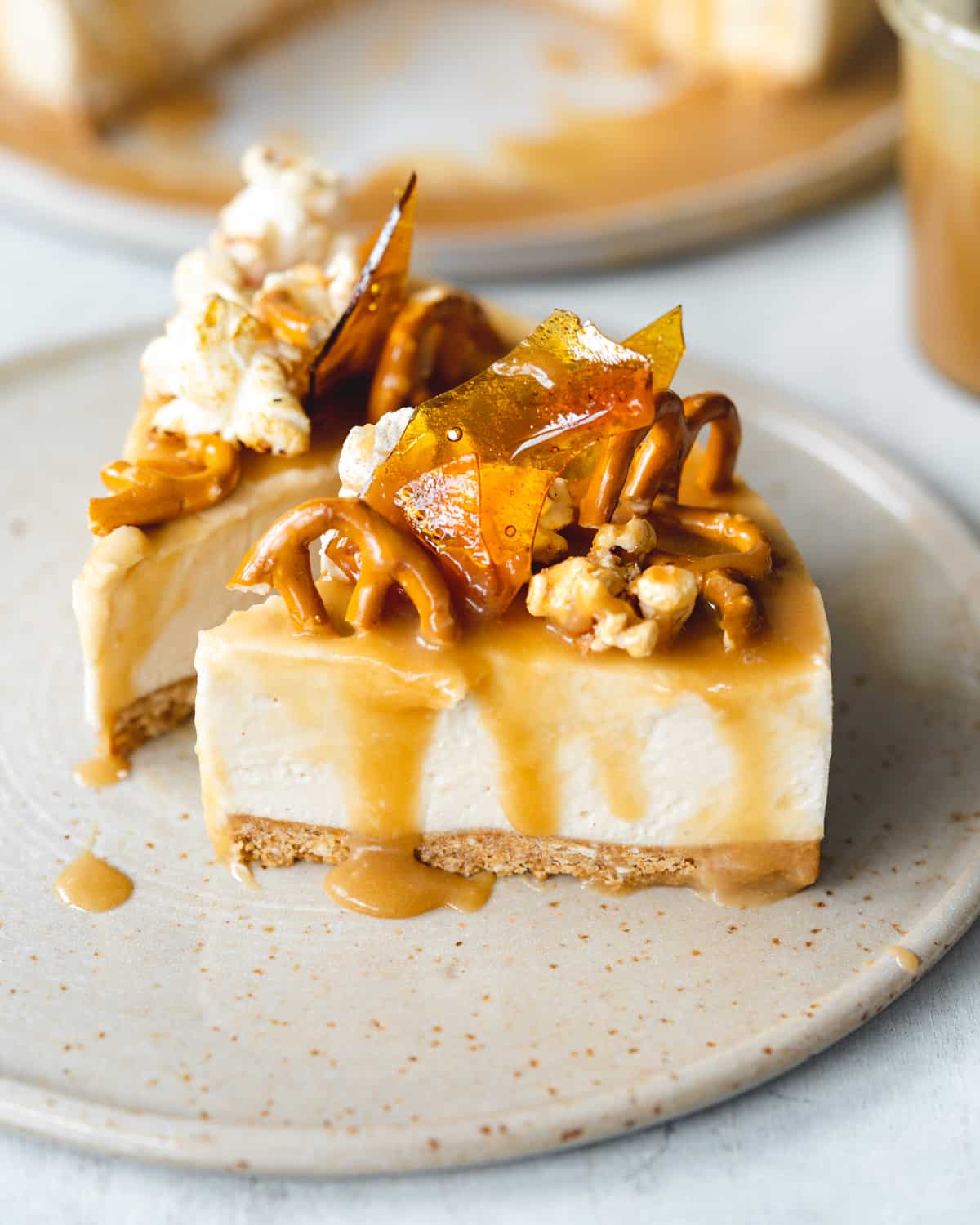 two slices of cheesecake with caramel sauce, pretzels, popcorn, and caramel shards on top, on a ceramic plate.