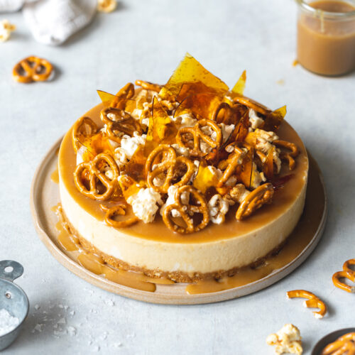 cheesecake topped with caramel sauce, pretzels, toffee popcorn, and caramel shards on a ceramic plate.