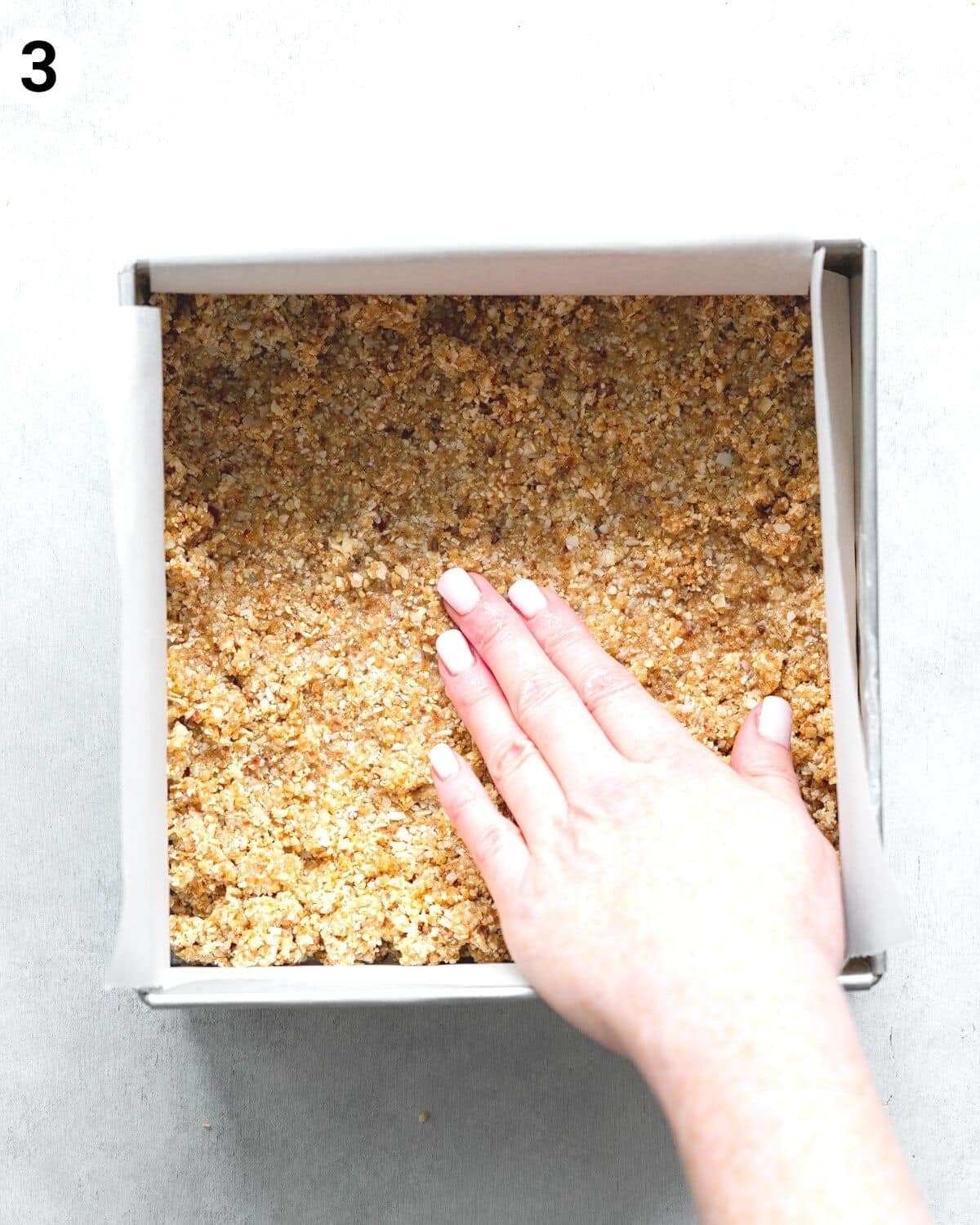 hand pressing cheesecake crust into a square pan lined with parchment paper.