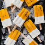 flatlay of coconut mango passion fruit popsicles on a bed of ice on black tray.