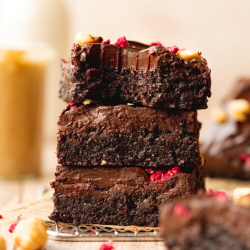 a stack of no-bake vegan brownies with ganache and freeze-dried raspberries on top, with a small jar of hazelnut butter in the background.