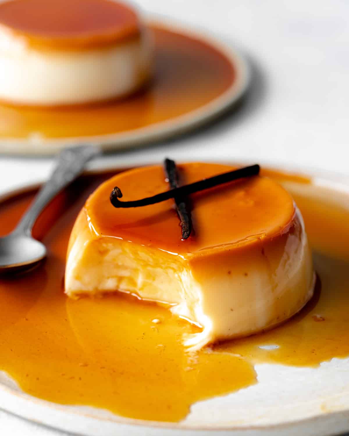 caramel flan on a plate with caramel sauce and vanilla pods on top and spoonful taken out of it.