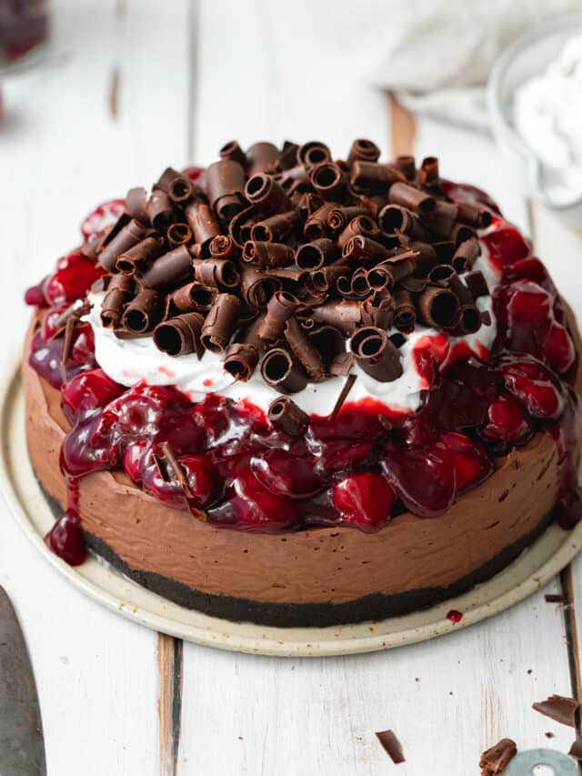 chocolate cheesecake with cherries, cream and chocolate curls on a white wooden surface.