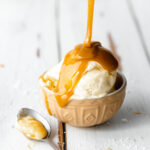 small bowl with vanilla ice cream and caramel sauce pouring over it, on a white wooden surface.