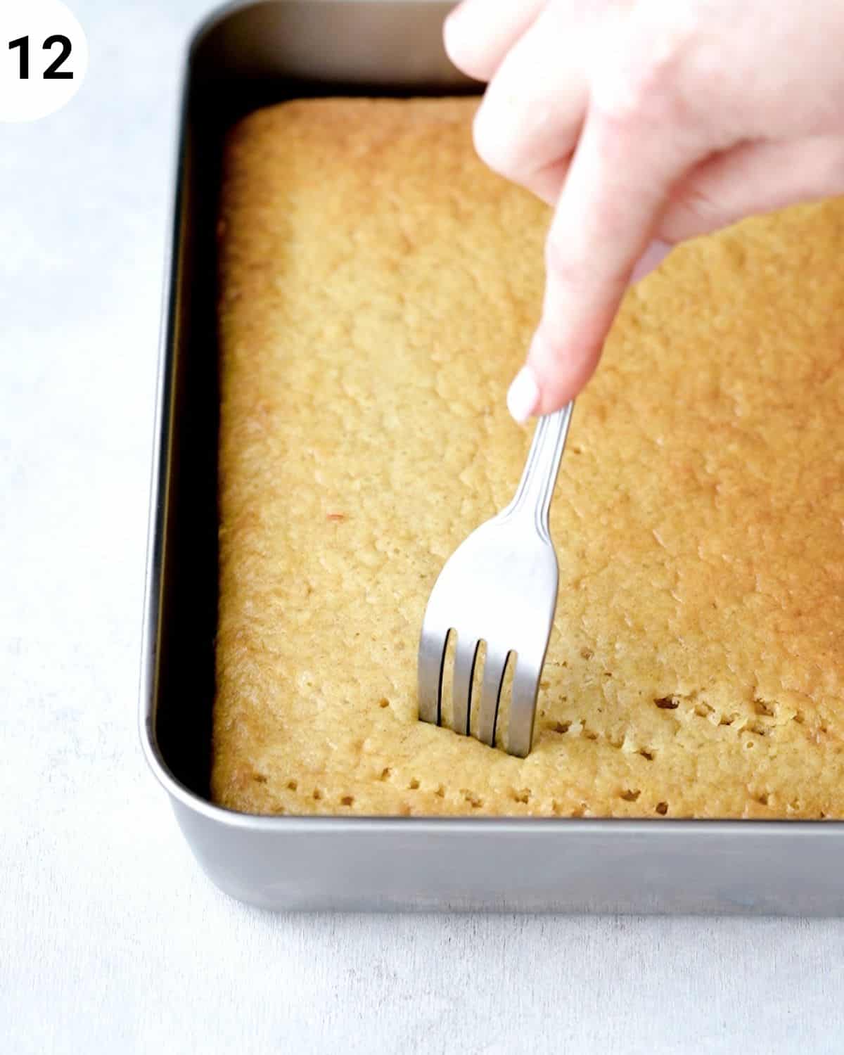 side view of tres leches cake in a metal square baking tin with fork piercing holes into it.