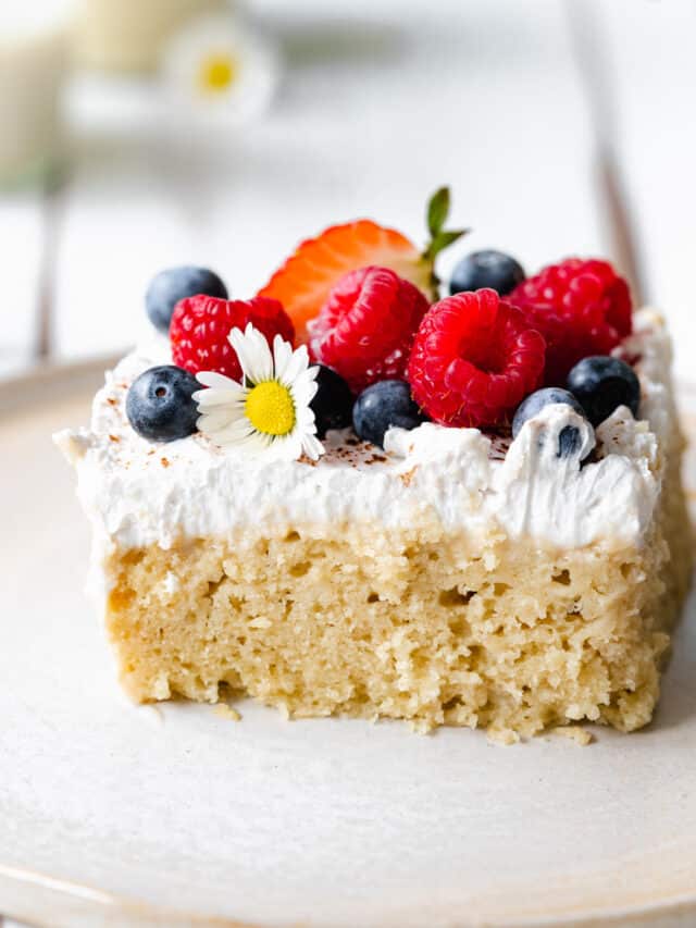 slice of vegan tres leches cake with fresh berries and daisies on top on a ceramic plate.