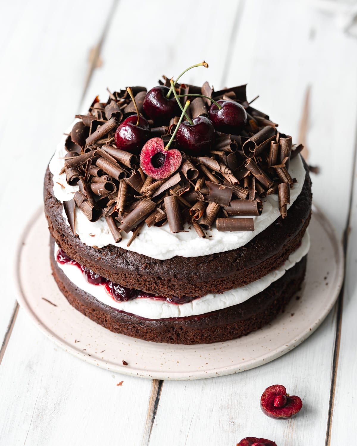 layered black forest cake with chocolate curls and cherries on top, on a white wooden surface.