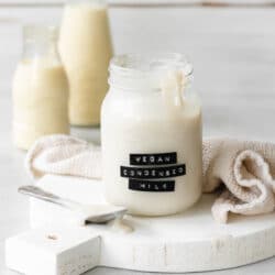 a labelled jar of vegan sweetened condensed milk on a white wooden tray with milk bottles in the background.