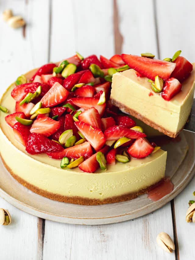 pistachio cheesecake topped with fresh strawberries and pistachio slivers on a white wooden surface.