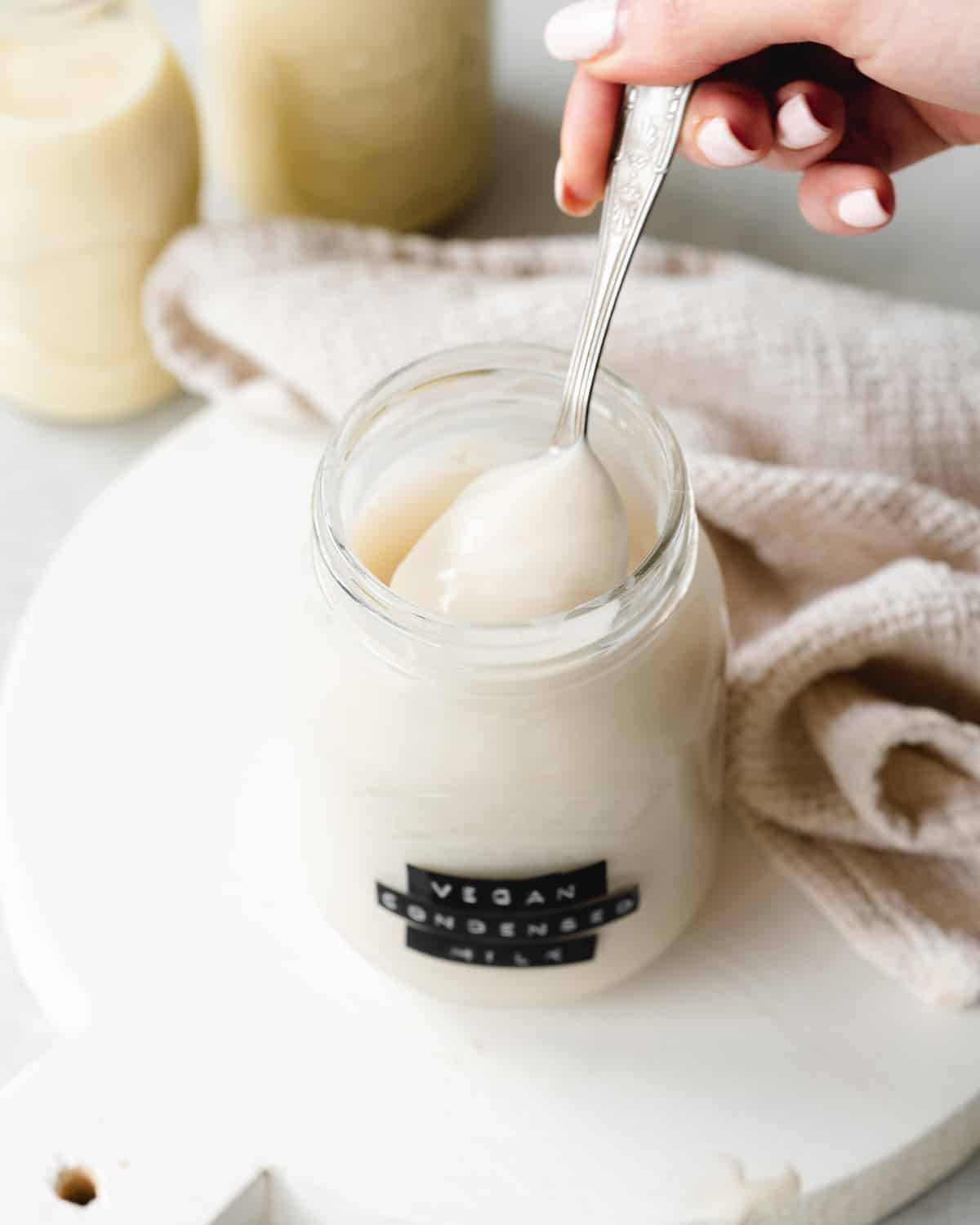 a labelled jar of vegan sweetened condensed milk on a white wooden tray with milk bottles in the background.