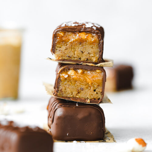 stack of chocolate peanut butter caramel bars with a jar of peanut butter in the background.