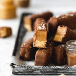 stack of homemade vegan twix bars on a vintage metal tray with a small jar or peanut butter in the background.