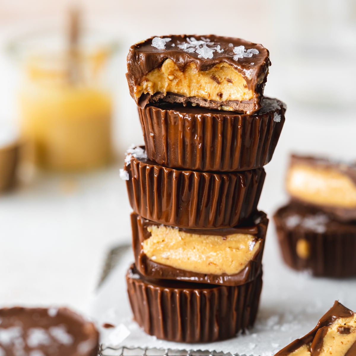 stack of homemade reese's cups on a wire rack with parchment paper and a jar of peanut butter in the background.