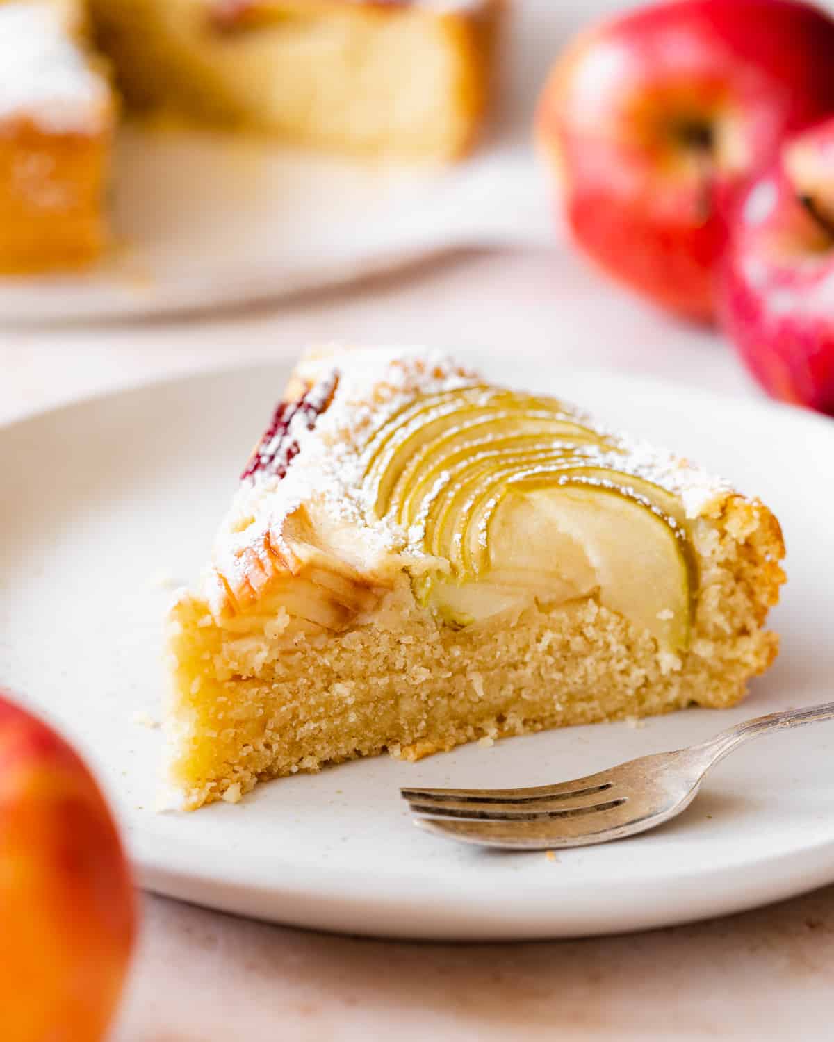 slice of cake with apple topping on a plate with a fork next to it, and red apples scattered around.