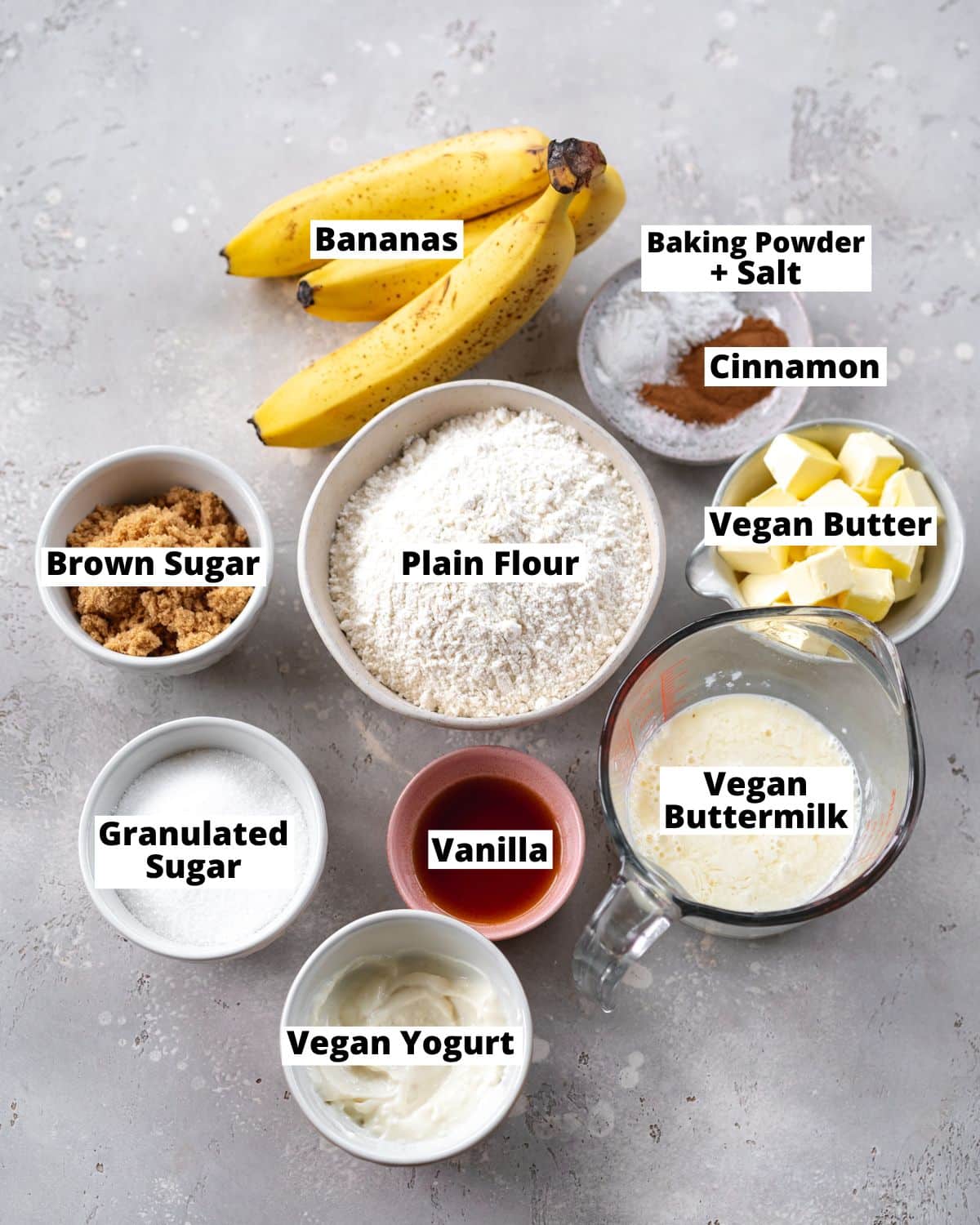 ingredients for vegan banana cake measured out in bowls on a grey stone surface.