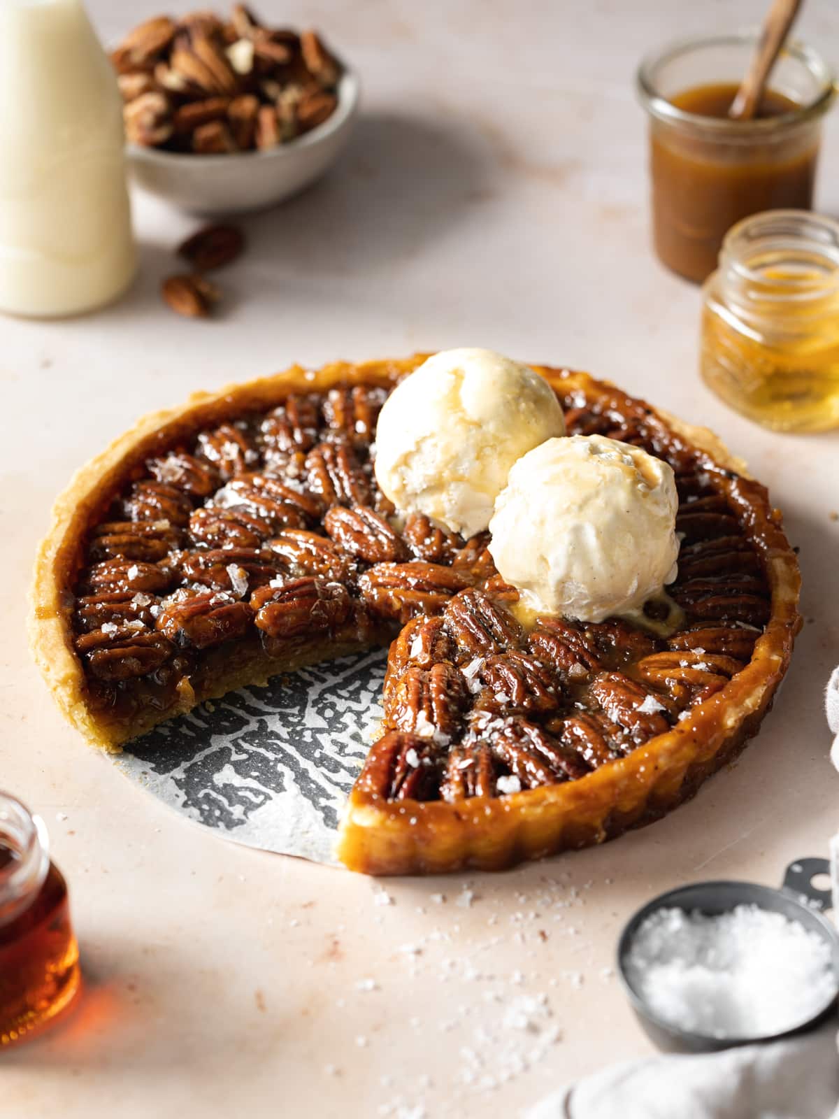 pecan pie with two scoops of vanilla ice cream and maple syrup on a peach marble surface.