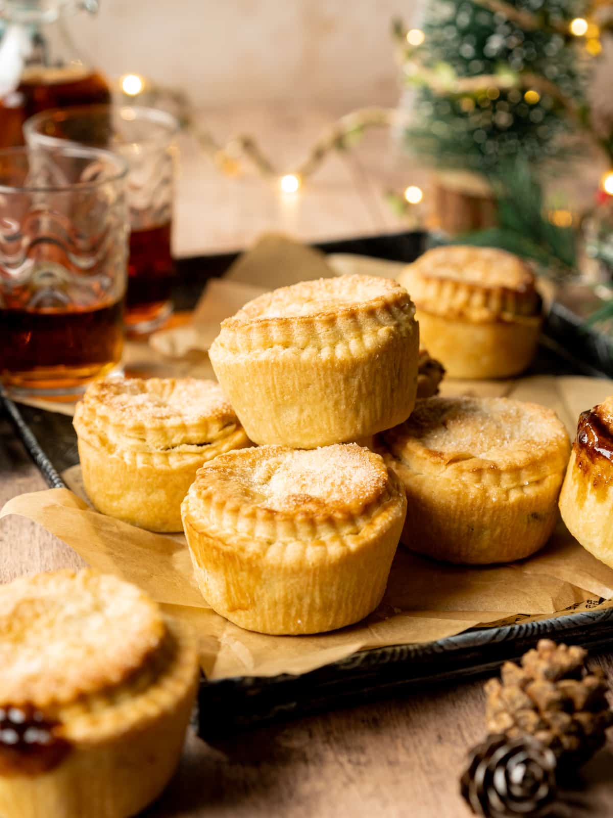 a stack of mince pies on a tray with a small christmas tree and bottle of brandy in the background.