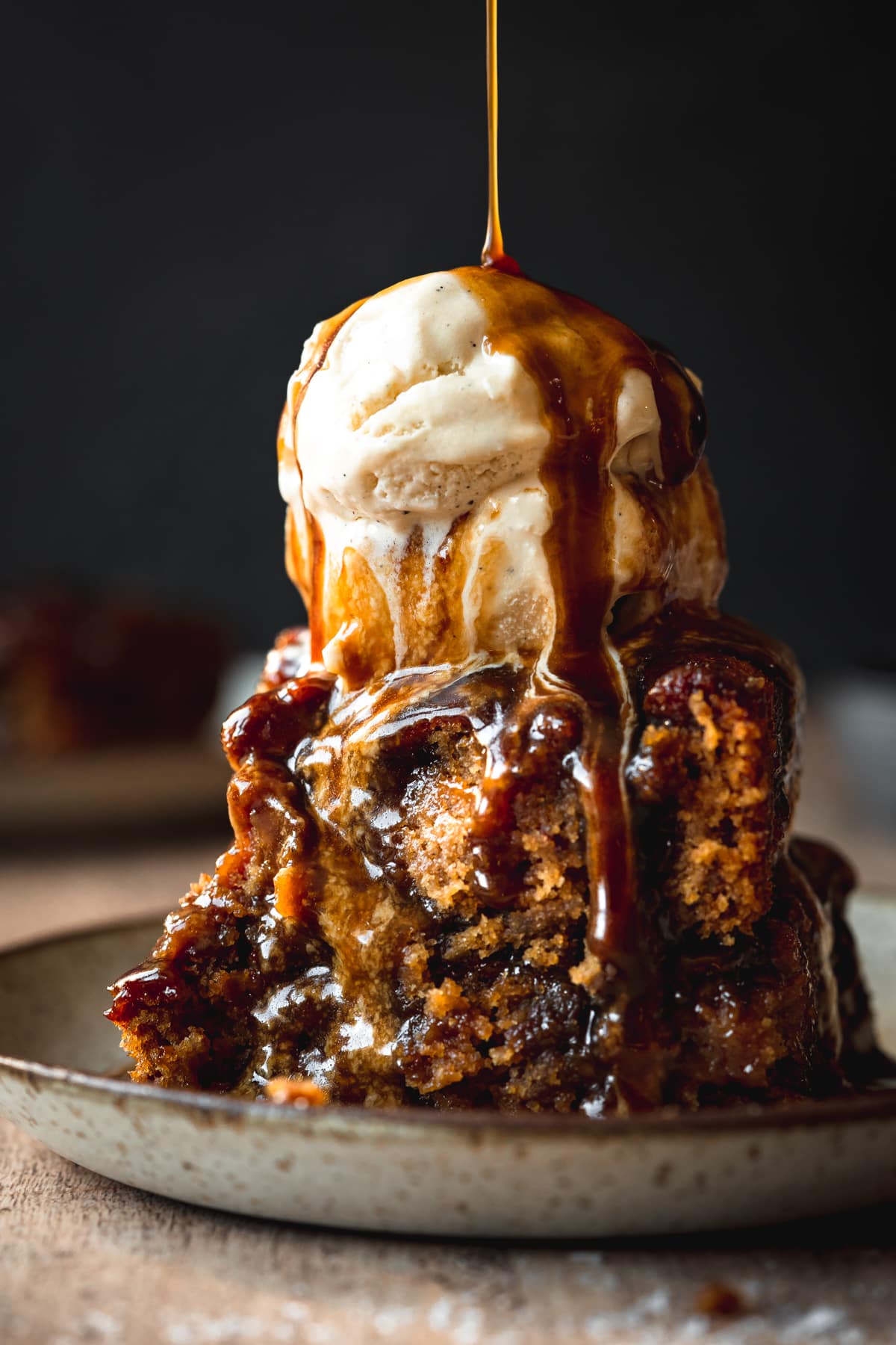 sticky toffee pudding on a ceramic plates with a scoop of vanilla ice cream on top, and caramel sauce poured over the top of it.