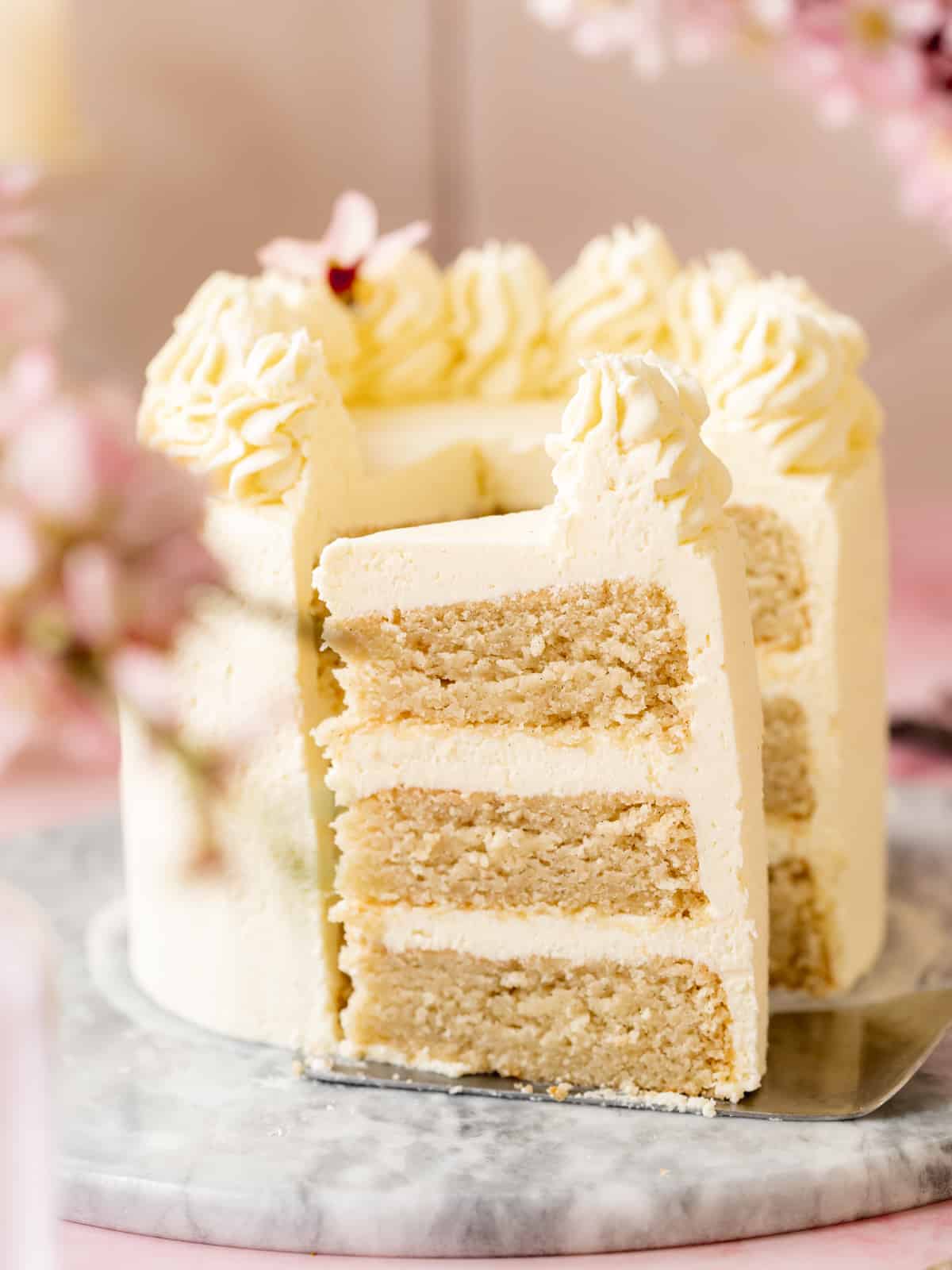 Side-on shot of 3-layer vegan vanilla cake with vanilla buttercream frosting with one slice removed and turned sideways on a cake server to show the distinct layers.