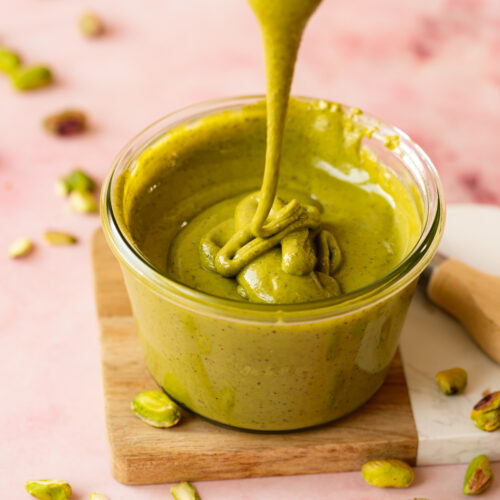 pistachio butter in a glass jar with some drizzling on top showing the smooth, runny, glossy consistency.