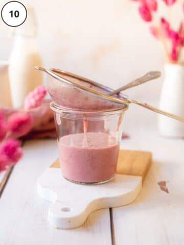 pink strawberry sauce passing through a small sieve into a glass jar with pink flowers in the background and foreground.