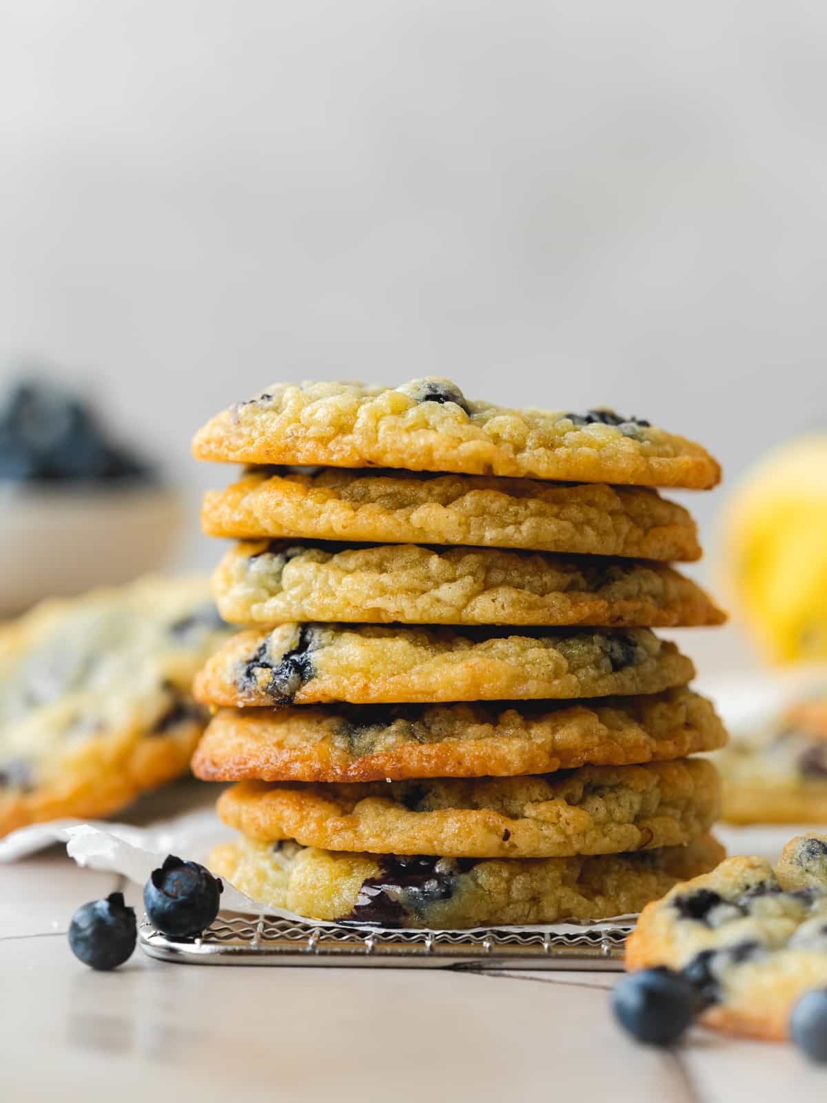 Side on shot of a stack of vegan lemon blueberry cookies on a piece of parchment on top of a cooling rack with a blurred out bowl of blueberries and a whole lemon in the background.
