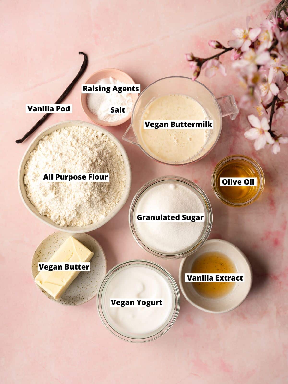 Ingredients needed to make vegan vanilla cake measured out into bowls on a pink table with a spray of cherry blossoms.