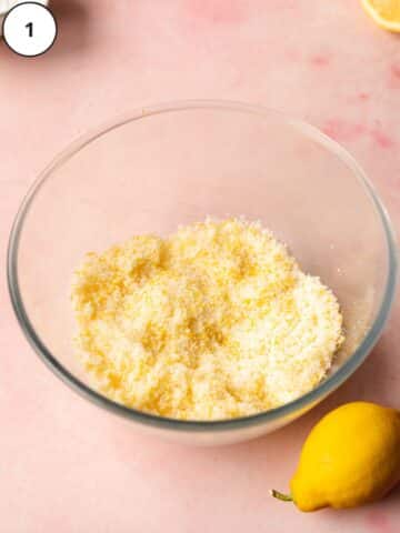 cane sugar with lemon zest rubbed into it in a large clear bowl. There are fresh lemons scattered around the bowl.