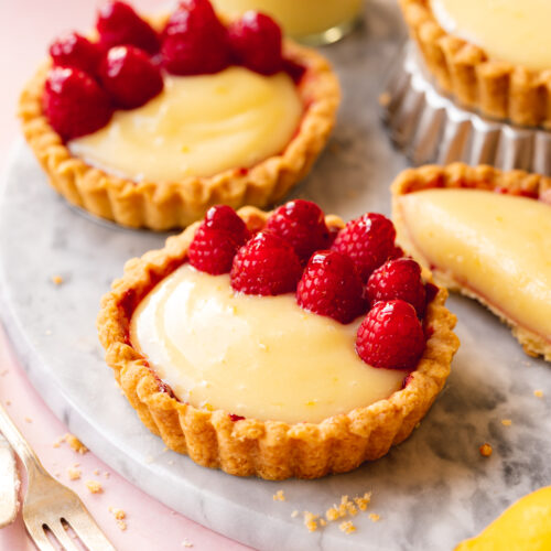 45 degree angle shot of vegan mini lemon tarts topped with raspberries on a marble serving tray.