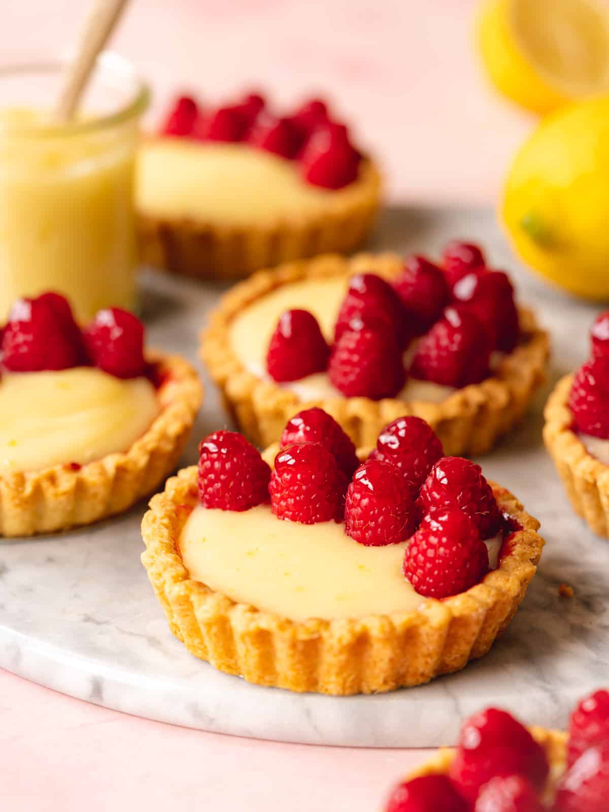 45 degree angle shot of several vegan lemon mini tarts topped with raspberries on a marble serving tray with a glass jar of lemon curd with a wooden spoon in it and several whole lemons in the background.