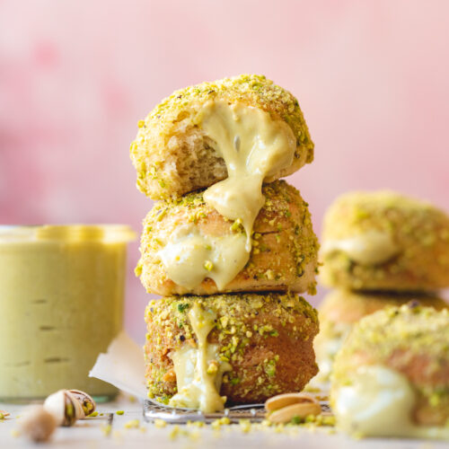 a stack of vegan donuts rolled in pistachio sugar and filled with pistachio cream on a wire rack. The background is contrasting pink and there are loose pistachios around the stack of donuts.