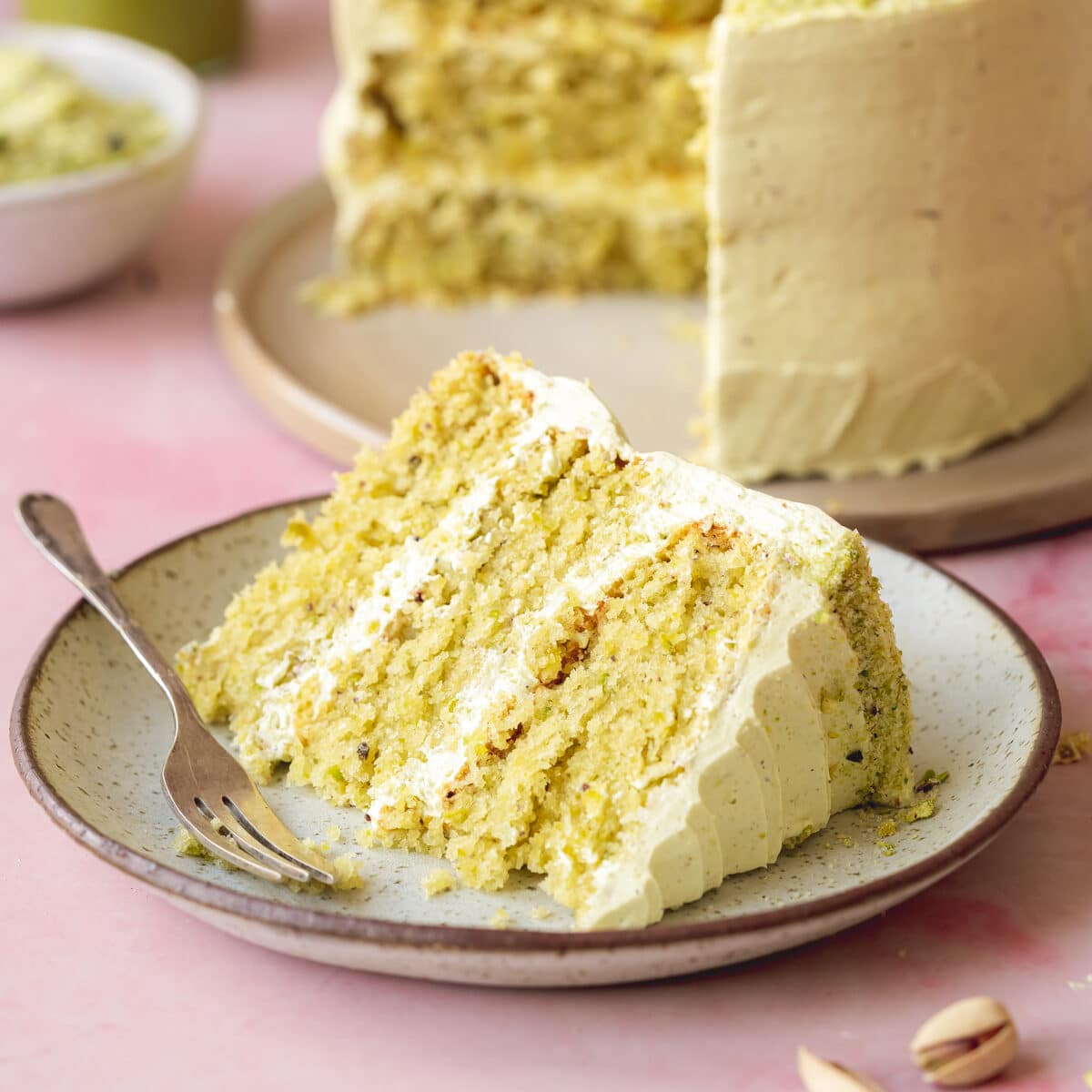 a slice of vegan pistachio cake layered with pistachio buttercream on a ceramic plate. There is a full cake in the background along with a small bowl of chopped pistachios.