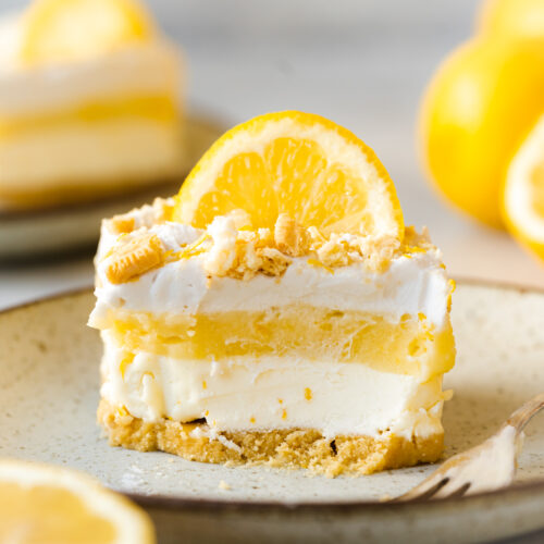 side on hero shot of a slice of lemon lush dessert lasagna on a plate showing the 4 clear layers of cookie crust, lemon cream cheese mousse, lemon curd, and whipped coconut cream topped with lemon zest, crushed cookies and a slice of lemon.