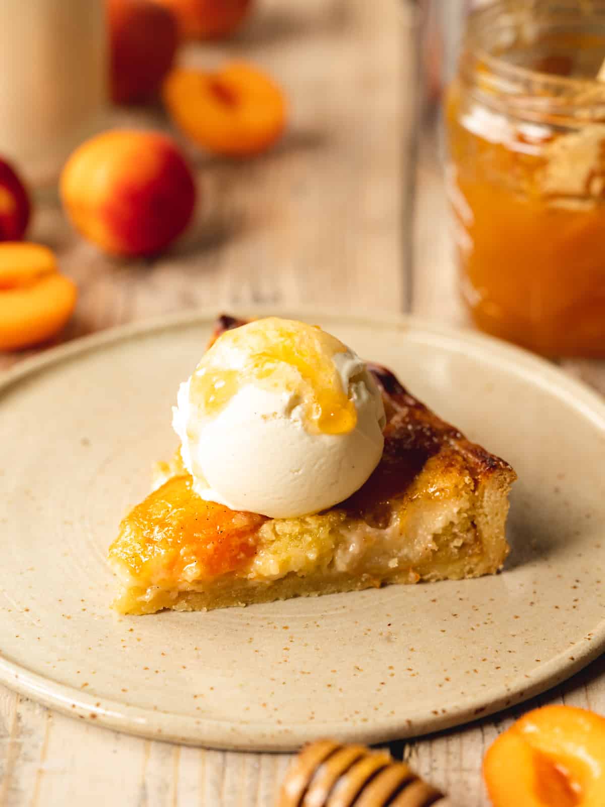 Slice of cooled apricot frangipane tart on an earthenware plate garnished with a scoop of vanilla vegan ice cream and an extra dollop of jam.