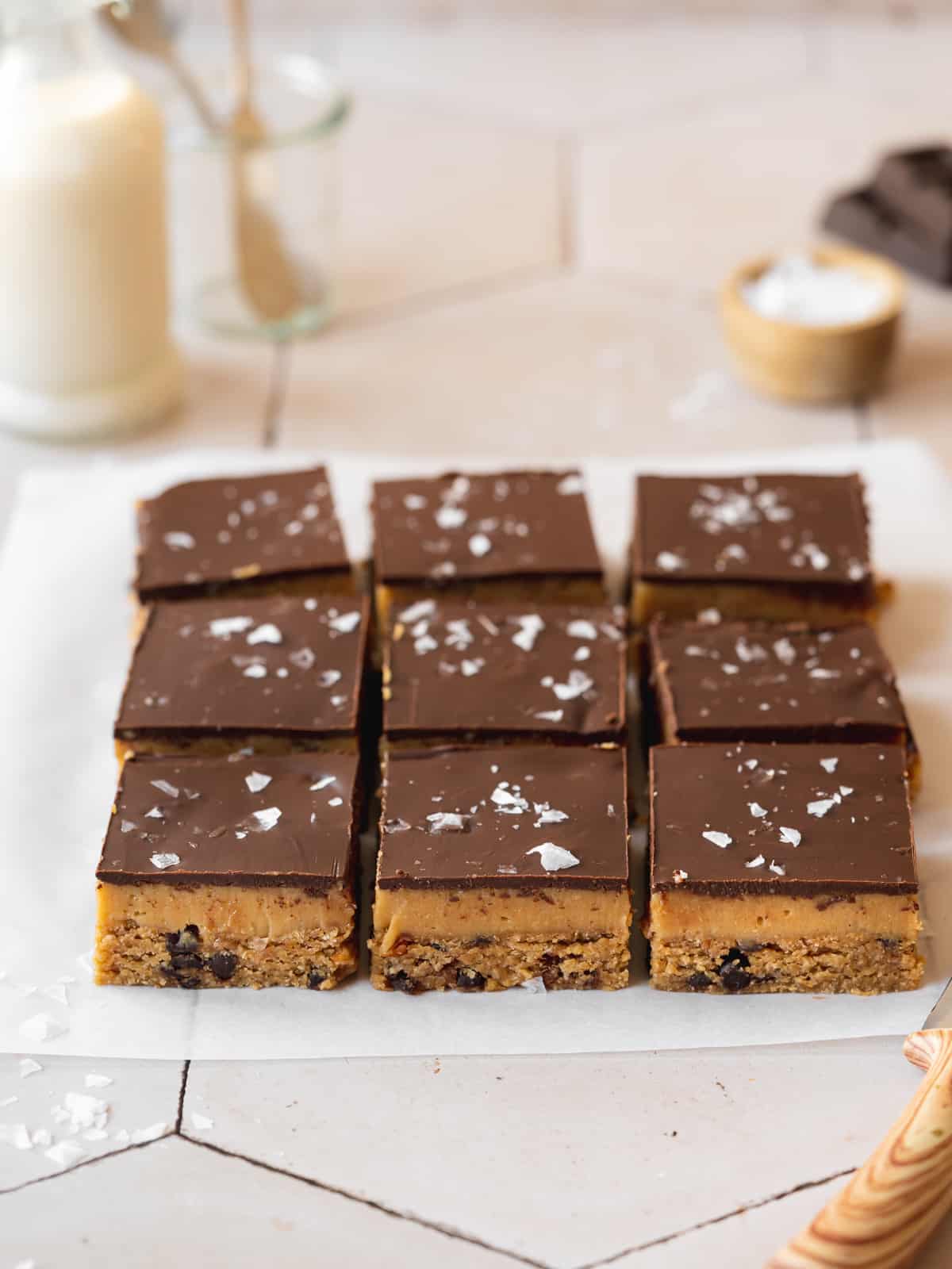 slices of vegan caramel cookie dough bars lined up on a sheet of parchment paper with a small jug of oat milk in the background.