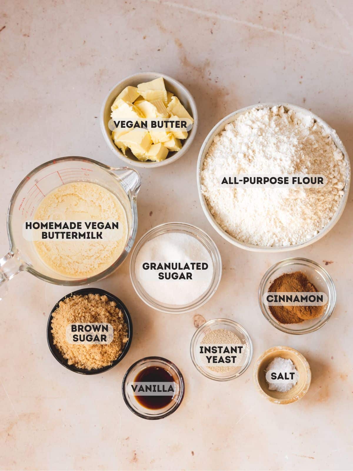 Ingredients needed to make vegan cinnamon buns measured out into bowls on a white table with text overlay.