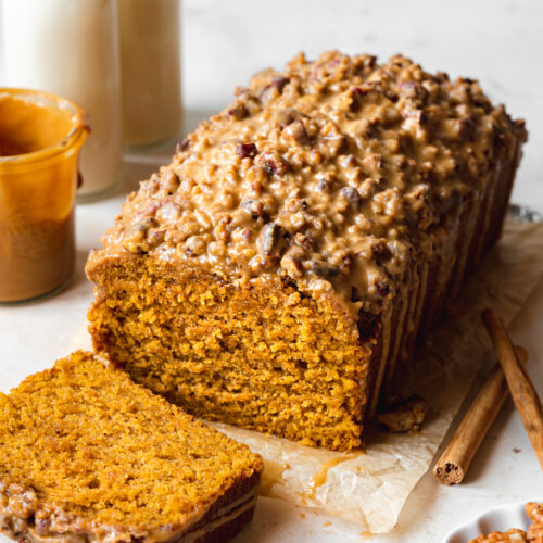 a loaf of vegan pumpkin bread with a slice taken from it showing the fluffy texture, It has a biscoff glaze and pecan streusel topping.