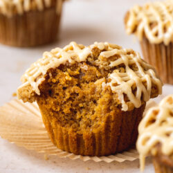 Closeup shot of a vegan pumpkin muffin topped with streusel and a hatchmark pattern of maple glaze with a bite taken out.