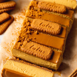 45 degree angle shot of no-bake biscoff pumpkin cheesecake bars with one tipped on its side to show the biscoff cookie crust, pumpkin cheesecake filling, and biscoff cookie spread topping.