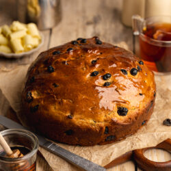 a round loaf of barmbrack bread on a bread board with crunched up brown paper and vegan butter, milk, and tea in the background.