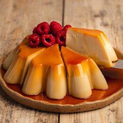 a large fluted flan with caramel sauce dripping down the sides on an earthenware plate, there are raspberries on top and a slice is being lifted away with a cake server showing the creamy consistency.