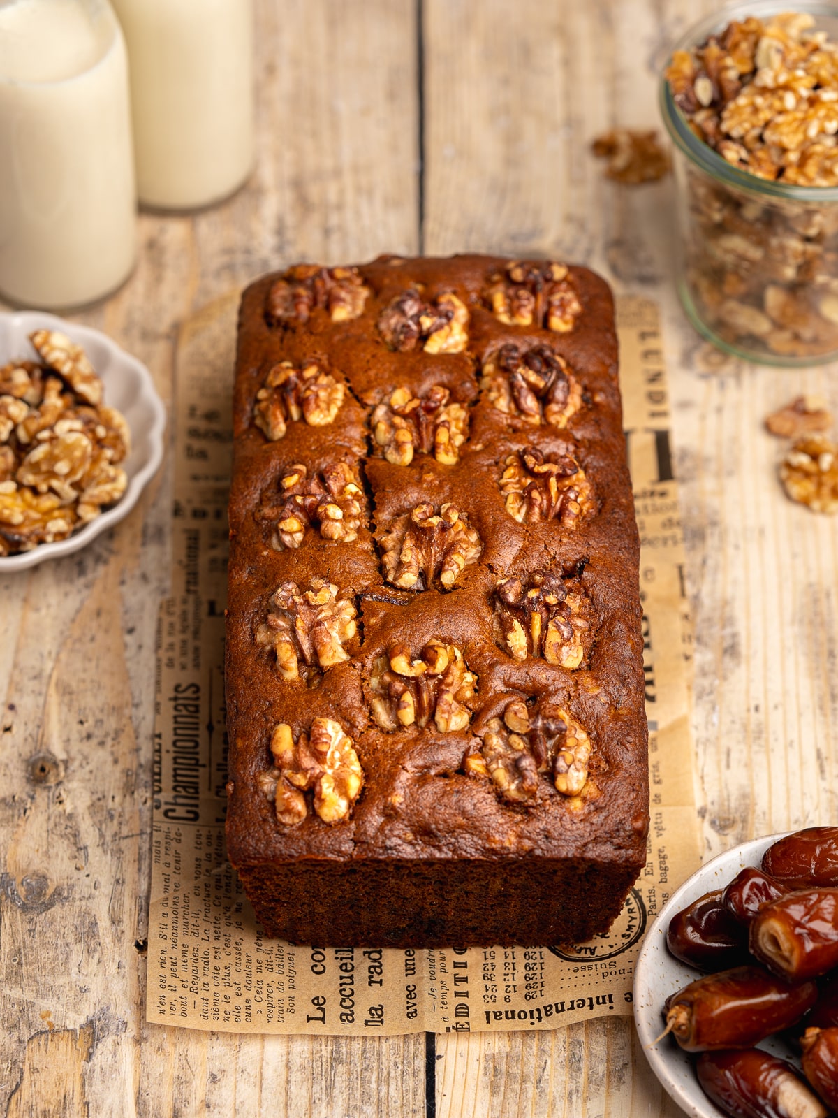 a loaf of freshly baked date cake with walnuts on top on a wooden surface with whole dates and walnuts scattered around.
