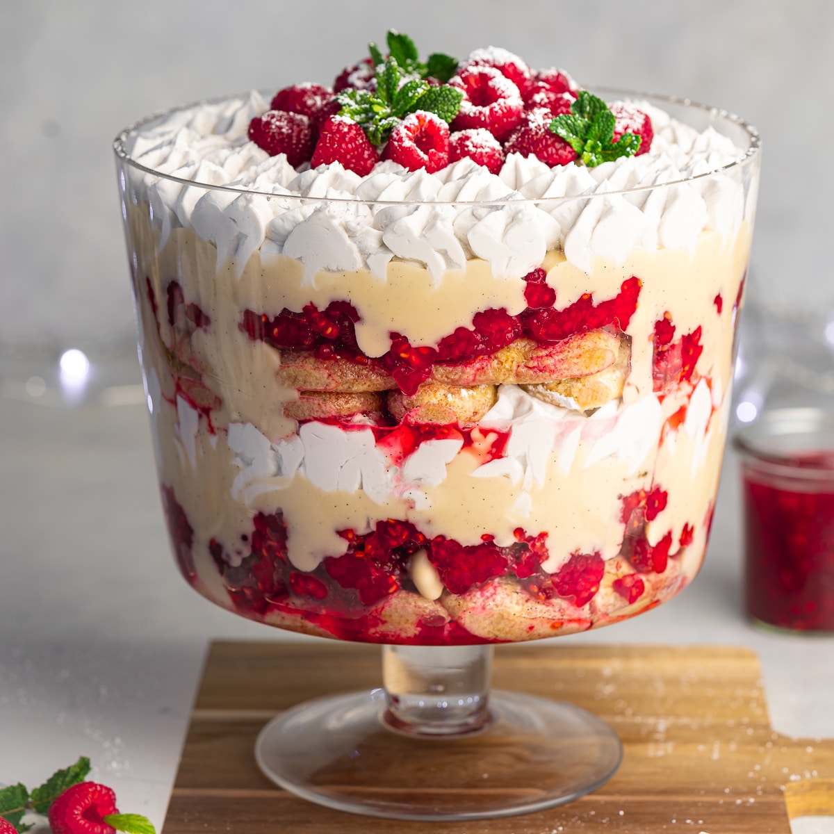 a trifle jar with layers of homemade ladyfingers, macerated raspberries, coconut white chocolate custard, and vegan whipped cream topped with raspberries and fresh mint leaves.