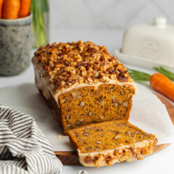 a loaf of carrot cake with brown butter glaze and chopped walnut topping on a wooden cutting board with fresh carrots in the background.