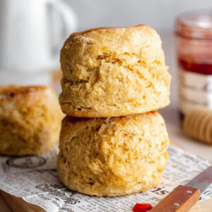 two freshly baked scones stack on top of one another on a sheet of newspaper with a jar of bonne maman strawberry jar in the background.
