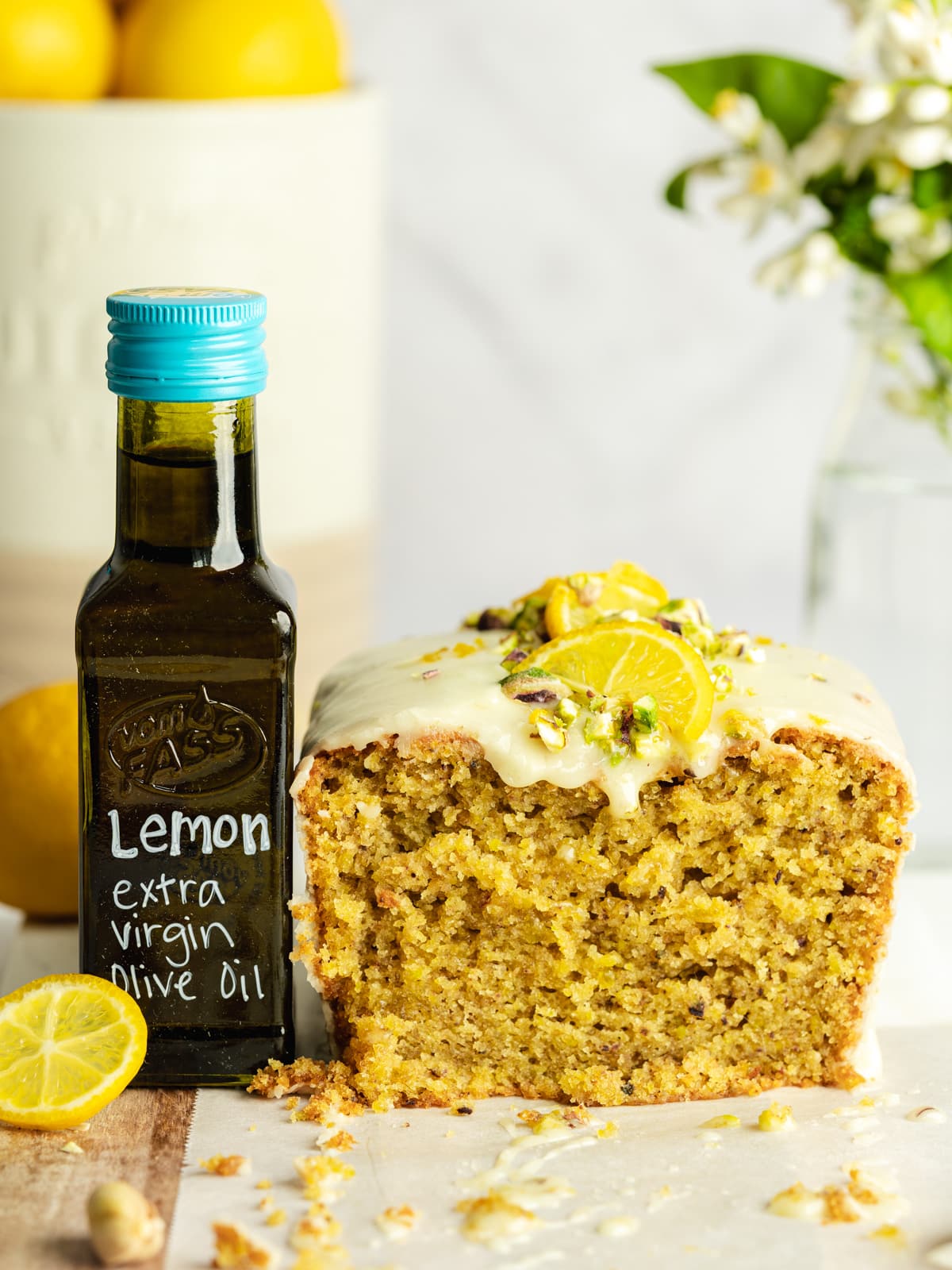 a lemon pistachio loaf with cream cheesecake glaze sliced open on a cutting board next to a small bottle of lemon extra virgin olive oil.