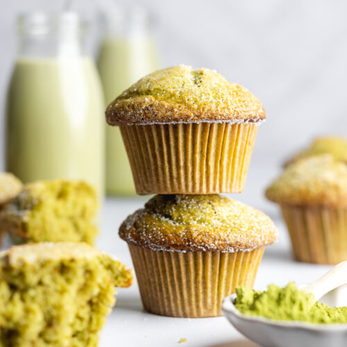 two matcha muffins stacked on top of each other with bottle of matcha milk in the background and a small pot of matcha tea powder in the foreground.