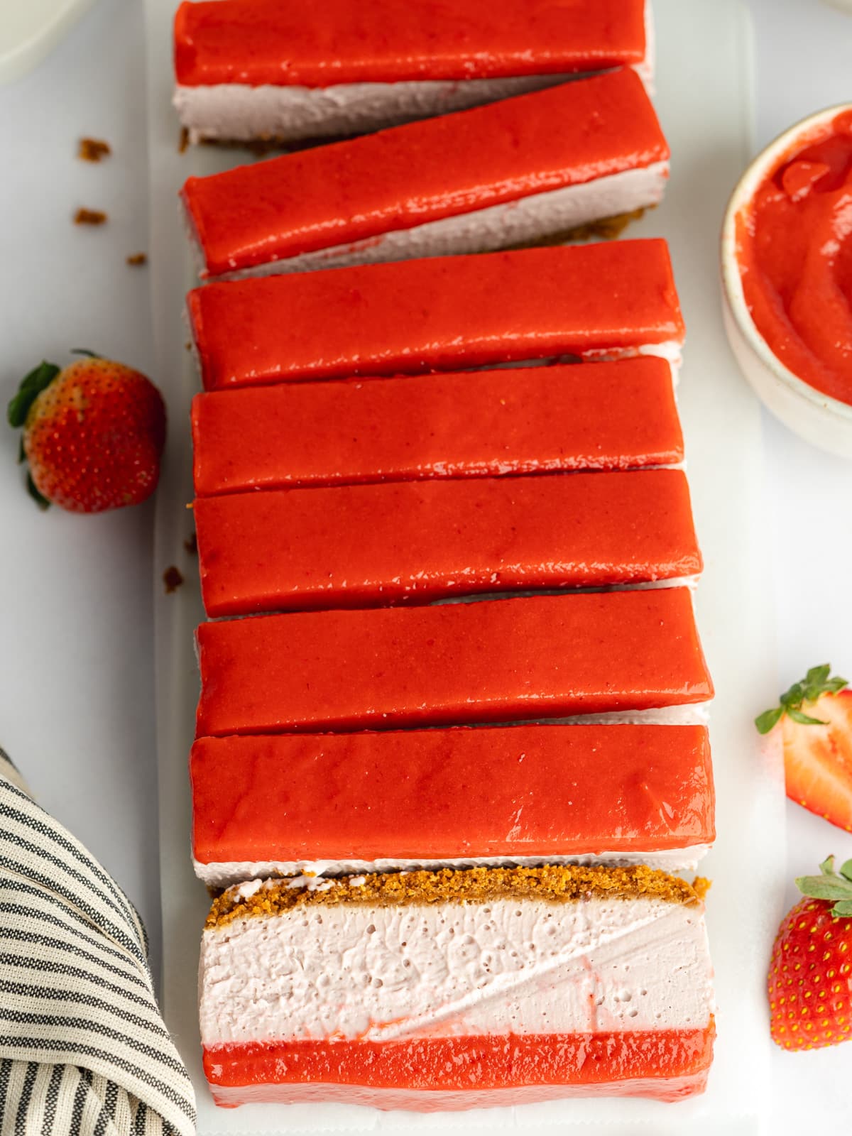 slices of strawberry cheesecake bars topped with strawberry curd and fresh strawberries scattered around.
