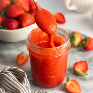 a glass jar of strawberry curd with a spoon lifting out some of the mixture showing the thick and smooth consistency.